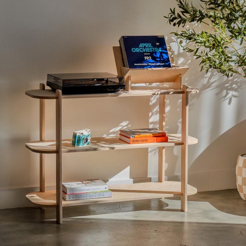 May include: A light-colored wooden record player stand with three shelves. The top shelf holds a black record player and a record holder with a record that says "April Orchestra FR2". The middle shelf has books and a small camera. The bottom shelf has books.