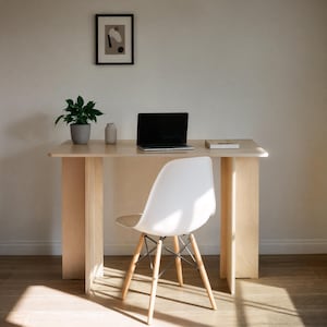 May include: A light wood desk with a laptop, book, and small vase. A white chair with wooden legs sits in front of the desk. A small potted plant and framed artwork are also visible. The desk is a light natural wood color.