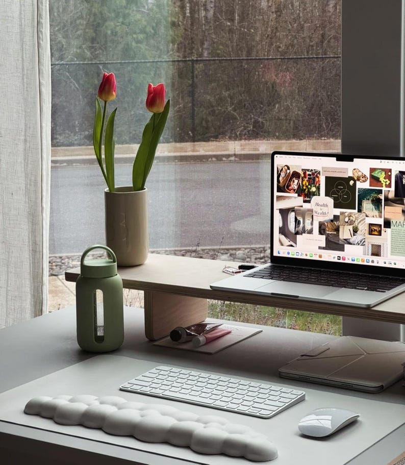 May include: A white laptop computer with a motivational collage on the screen is on a wooden stand. A green glass water bottle, a white keyboard, and a white mouse are on a gray desk mat. A vase with two pink tulips is on the stand.