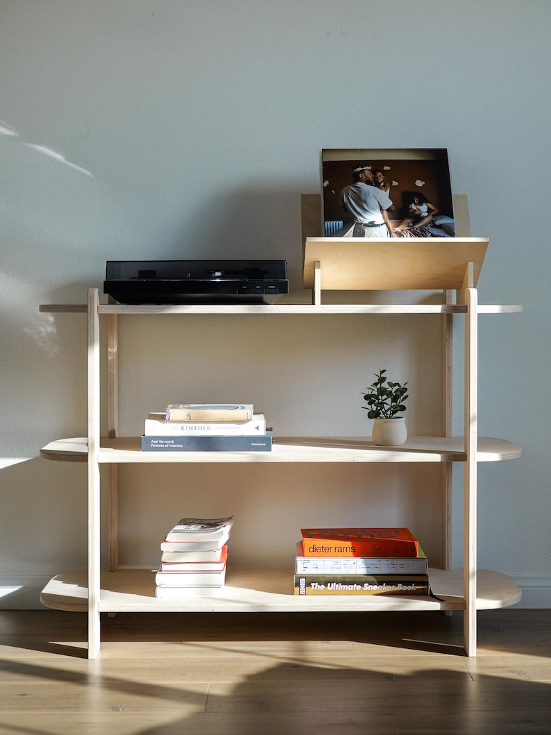 May include: A light-colored wooden shelving unit with three shelves. A black record player sits on the top shelf, with a record and stand next to it. Books and a small potted plant are on the other shelves.