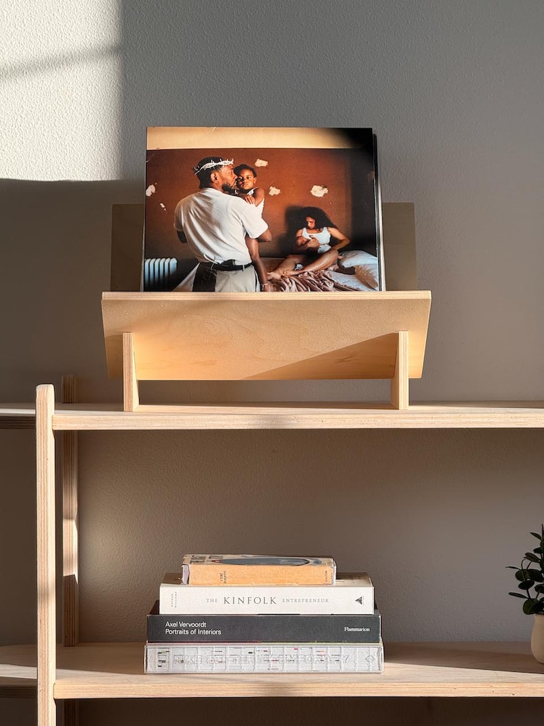May include: A wooden shelf unit with a framed photograph of a family. The photograph is displayed on a wooden stand. Below the stand are several books, including "The Kinfolk Entrepreneur". The shelf is made of light-colored wood.