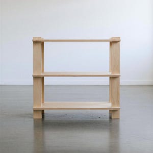 May include: A light-colored wooden shelving unit featuring three horizontal shelves supported by four vertical posts. The structure is composed of smooth, unfinished light wood panels with visible grain, creating a simple, open-frame design.