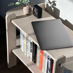 May include: A light-colored wooden bookshelf with two shelves. A laptop, a black alarm clock, and a small vase with a plant are on the top shelf. Books are neatly arranged on the lower shelf.