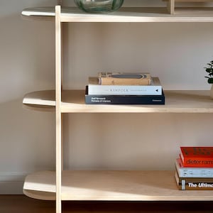 May include: A light wood bookshelf with three shelves. The top shelf is empty. The middle shelf has a stack of four books, including "Kinfolk: The Entrepreneur". The bottom shelf has a stack of three books, including "Dieter Rams" and "The Ultimate Guide to Interior Design".