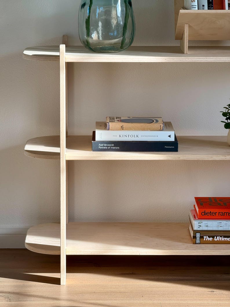 May include: A light wood bookshelf with three shelves. The top shelf is empty. The middle shelf has a stack of four books, including "Kinfolk: The Entrepreneur". The bottom shelf has a stack of three books, including "Dieter Rams" and "The Ultimate Guide to Interior Design".