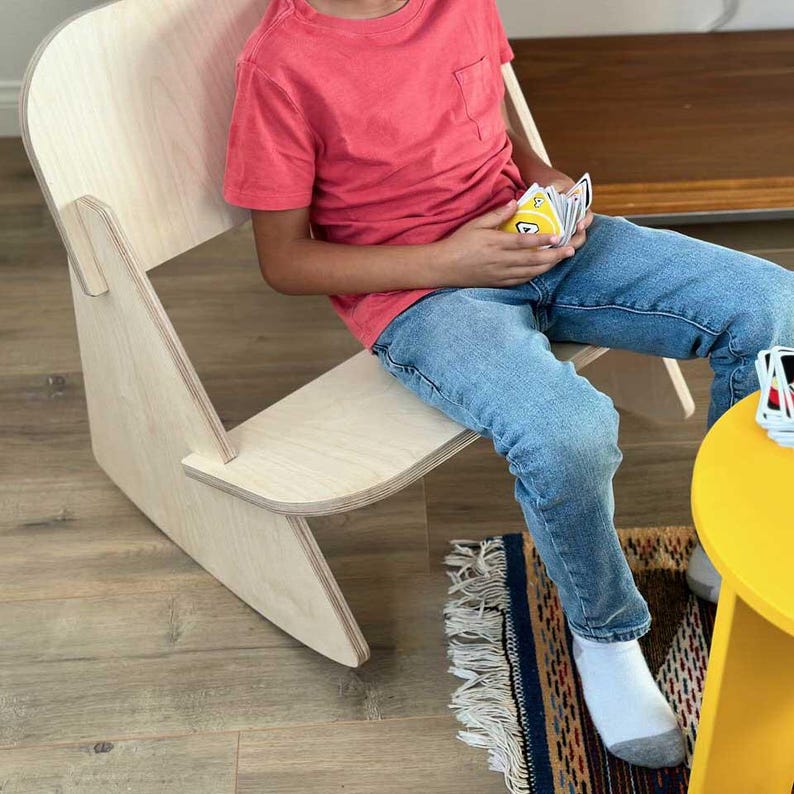 May include: A child sits on a light wood rocking chair with a yellow table and rug in the background. The child is holding a deck of cards.