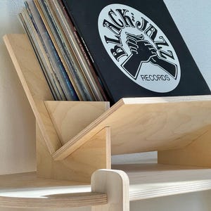 May include: A wooden record holder with a slanted design, holding several vinyl records. The top record is black with white text and a graphic that reads "BLACK JAZZ RECORDS". The holder is made of light-colored wood and sits on a shelf.