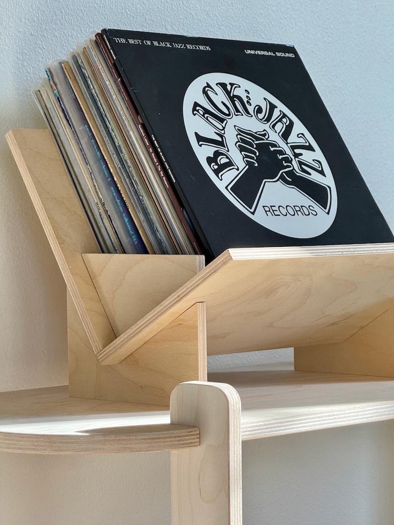 May include: A wooden record holder with a slanted design, holding several vinyl records. The top record is black with white text and a graphic that reads "BLACK JAZZ RECORDS". The holder is made of light-colored wood and sits on a shelf.