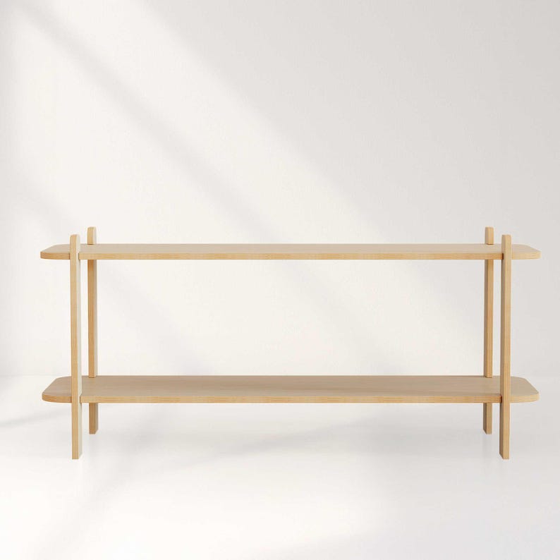 May include: A light-colored, two-tiered wooden shelf with a minimalist design. The shelf has two horizontal planks supported by vertical posts at each end, creating a simple, modern aesthetic. The wood has a natural finish.