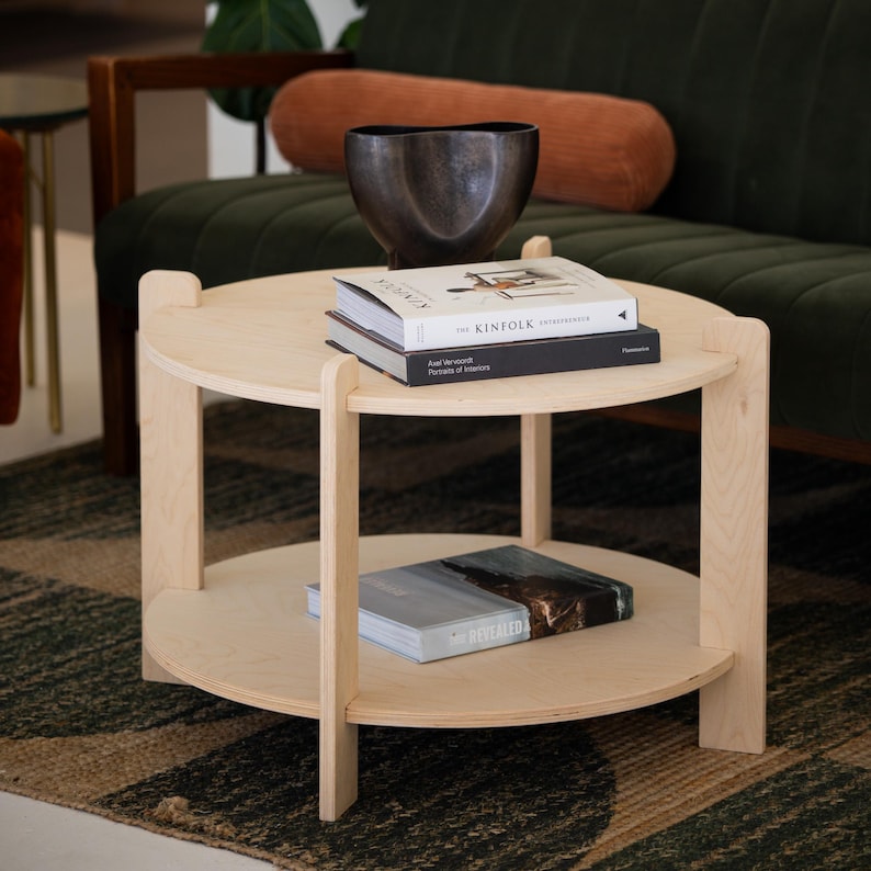 Peut inclure: Une table basse ronde &agrave; deux niveaux en bois clair, avec une finition naturelle. L'&eacute;tag&egrave;re sup&eacute;rieure contient deux livres et un bol en m&eacute;tal fonc&eacute;. L'&eacute;tag&egrave;re inf&eacute;rieure a un seul livre. La table est devant un canap&eacute; vert.
