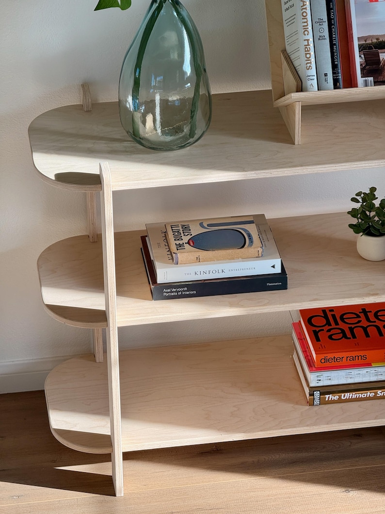 May include: A light wood bookshelf with three shelves. The shelves are rounded at the front. The top shelf has a clear glass vase with a green plant. The middle shelf has a stack of books, including "The Bugatti Story" and "Kinfolk". The bottom shelf has a stack of books with the title "Dieter Rams".