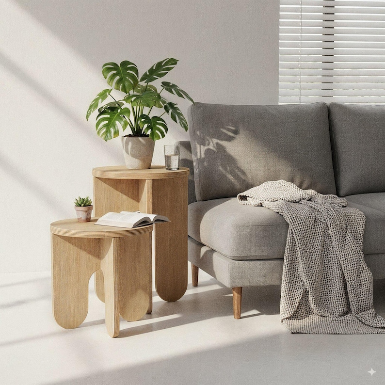 May include: Two light wood side tables of different heights. A potted plant with large green leaves sits on the taller table. A small succulent in a terracotta pot and an open book are on the shorter table. A gray sofa and blanket are in the background.