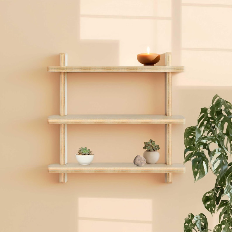May include: A three-tiered wooden shelf with a light wood finish. The shelf is mounted to a light peach wall. There are two small potted plants and a candle on the shelf.