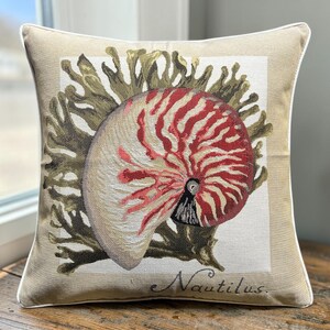 May include: A square throw pillow with a beige background and white piping. The centre features a woven tapestry design of a nautilus shell with red and white stripes, set against green coral. The word Nautilus is printed in black script.