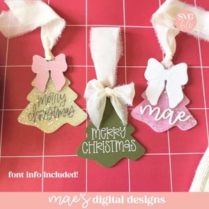 May include: Three Christmas tree ornaments with white ribbon and bows. One tree is gold with "Merry Christmas" in script, another is green with the same text, and the last is pink with the name "mae".