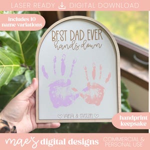 Puede incluir: Un recuerdo de huellas de manos de madera con el texto "BEST DAD EVER hands down". El diseño incluye dos huellas de manos en rosa y morado, con los nombres "Vania & Evelyn" debajo. También se ve el texto "handprint keepsake".