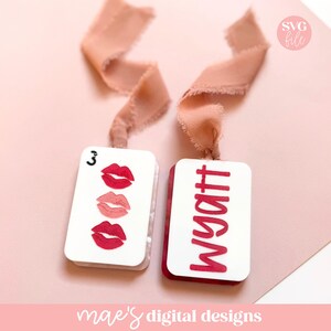 May include: Two white rectangular tags with red accents and pink ribbons. One tag features three red lip illustrations and the number 3. The other tag displays the name "Wyatt" in red. The background is a soft pink.