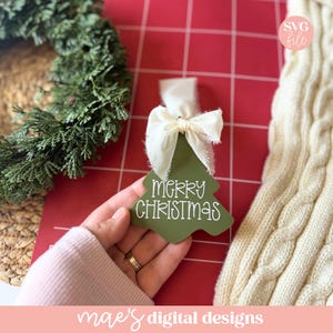 May include: A green Christmas tree-shaped tag with the words "MERRY CHRISTMAS" in white script. The tag is adorned with a white ribbon and bow. The image also includes a green wreath and a cream-colored knitted scarf.