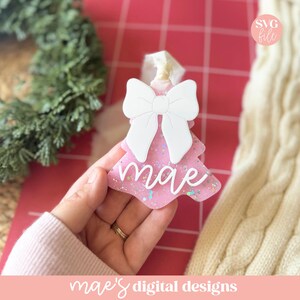 May include: Pink Christmas tree ornament with a white bow and the name "mae" in white script. The ornament has glitter accents and is held against a red background. The text "SVG file" is in the upper right corner.
