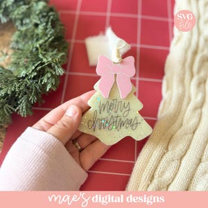 May include: A Christmas tree-shaped ornament with a pink bow at the top. The ornament is light green with glitter and the words "merry christmas" in black script. The ornament is held by a person.