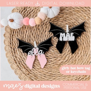 Op de afbeelding: Twee zwarte vleermuisvormige labels met witte letters "ella" en "MAE". De labels hebben roze strikaccenten en worden weergegeven op een geweven oppervlak. De tekst "girly bat bow tag or keychain" is ook zichtbaar.