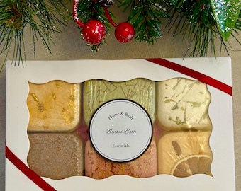 May include: A white rectangular gift box with a scalloped window containing six square bath bombs in various colors and textures. A central circular label reads Home & Bath Bonsai Bath Essentials. The box is decorated with a red ribbon.