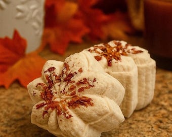 May include: Two off-white, flower-shaped bath bombs or soaps with a textured, molded surface. The top of each item is decorated with scattered, dried reddish-brown botanical petals embedded into the center of the floral design.