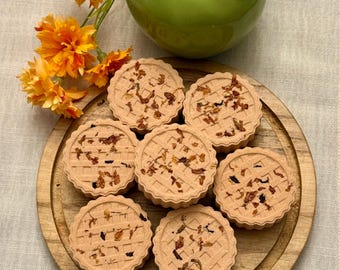 May include: A wooden serving tray holds seven round, light brown pastries with a lattice design and scattered dried petals. A green ceramic apple and orange flowers are in the background.