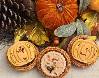 May include: Two orange pumpkin-shaped bath bombs and one round waffle-textured bath bomb, each nestled in a small woven wicker basket. The pumpkin bath bombs feature gold glitter accents and dried botanical pieces, while the waffle bath bomb has dried herbs.