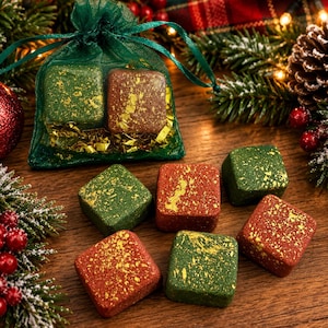 May include: A collection of square-shaped bath bombs in red and green colors, each featuring gold-colored flecks. Two bath bombs are contained within a sheer green drawstring pouch, while six others are arranged on a wooden surface.