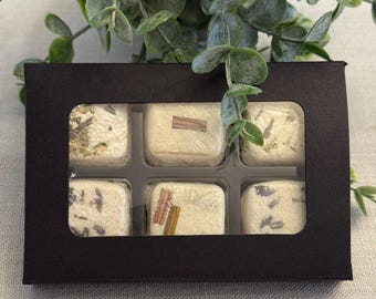 May include: A black box with a clear window containing six square, individually wrapped bath bombs. The bath bombs are off-white and decorated with herbs and spices. Green foliage is visible in the background.