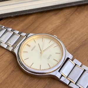 May include: A silver and gold Seiko Spirit wristwatch with a cream-coloured face. The watch has a rectangular link band and gold hands and markers. The word "Seiko" is printed at the top, and "spirit" is printed at the bottom of the watch face.