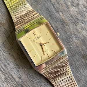 Könnte beinhalten: Eine goldfarbene Aureole Quartz-Uhr mit rechteckigem Zifferblatt und Mesh-Armband. Das Zifferblatt ist hellgoldfarben mit schwarzen Stundenmarkierungen. Die Zeiger sind schwarz und goldfarben. Die Uhr liegt auf einer Holzoberfläche.