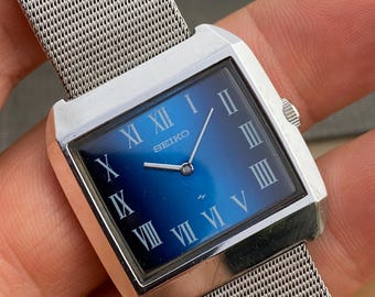 May include: A square Seiko wristwatch featuring a blue gradient dial with silver Roman numeral hour markers and silver hands. The watch has a polished silver-tone metal case and a flexible silver-tone stainless steel mesh strap.