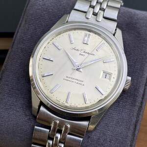 May include: A silver-toned Seiko Champion 860 wristwatch with a cream-colored dial, silver baton indices, and a date window at the three o'clock position. The watch features a stainless steel link bracelet and displays the text Seiko Champion 860.