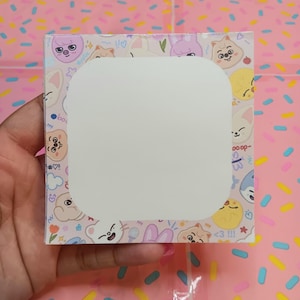 May include: A square notepad with a white speech bubble shape in the center. The notepad's border features a light pink background with cartoon animal faces and small icons. The notepad is held by a hand.