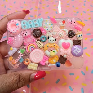 Op de afbeelding: Een rechthoekig doorzichtig plastic doosje versierd met een dichte verzameling kleurrijke miniatuur harsbedels, waaronder een lichtblauw BABY-bordje, een roze teddybeer, een roze donut, een regenboog en diverse snoepjes en hartjes.