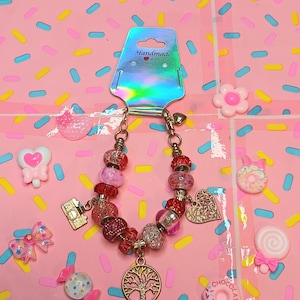 May include: A handmade charm bracelet with red, pink, and clear beads, gold-colored charms, and a tree of life pendant. The bracelet is displayed on a pink background with decorative elements. The tag reads "Handmade with love."