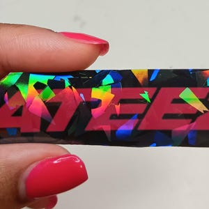 May include: A rectangular sticker with the word "ATEEZ" in red, set against a black background with a holographic, multicolored pattern. The sticker is held by a hand with red nail polish.