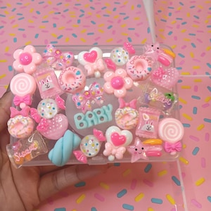 Puede incluir: Una funda transparente para teléfono decorada con adornos de caramelos rosas y blancos. La funda presenta varias formas, incluyendo flores, corazones y piruletas, con la palabra "BABY" en azul. El fondo es un patrón rosa.