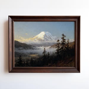 May include: A framed landscape painting depicts a snow-capped mountain rising above a valley filled with fog. Dark green trees line the foreground, and the painting is encased in a dark brown wooden frame.
