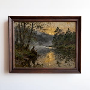 May include: A framed landscape painting depicts a person fishing on a riverbank at dusk. The artwork features a dark brown wooden frame, a serene river scene, and a misty atmosphere with trees and a golden sky.