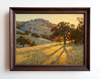 May include: A framed landscape painting depicts a sunlit hillside with trees and a rocky peak. The artwork features warm tones of gold, brown, and green, with long shadows cast by the trees. The frame is a dark brown color.