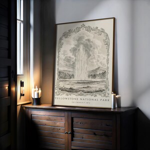 May include: A framed print of Yellowstone National Park, featuring a detailed black and white illustration of a geyser erupting. The print is displayed on a wooden cabinet, with candles nearby. The text "YELLOWSTONE NATIONAL PARK" is visible.
