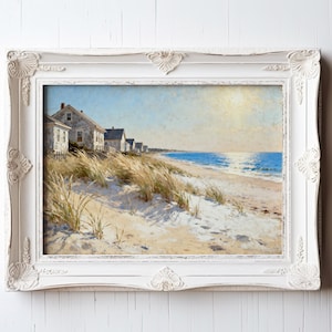 May include: A framed beach scene painting. The artwork depicts a row of weathered houses along a sandy beach with tall grasses in the foreground. The sky is a light blue, and the ocean is a deeper blue. The frame is white with ornate detailing.