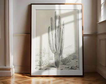 May include: A framed black and white botanical print of a saguaro cactus in a desert landscape. The artwork is displayed in a room with a wooden floor and white walls. The frame is a warm brown color.