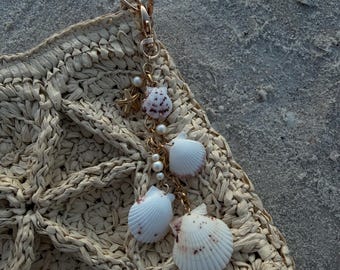 May include: A beige woven straw bag featuring a gold-toned metal lobster clasp charm. The charm is adorned with small white pearls, a miniature starfish, and four distinct white seashells with subtle brown speckles attached by gold chains.