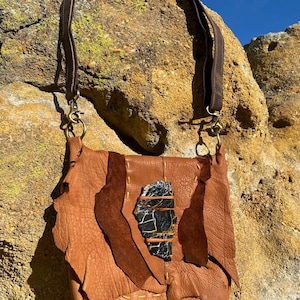 May include: A handmade brown leather shoulder bag with a dark brown strap and brass-colored hardware. The bag features a large, irregularly shaped black and white stone accent on the front. The bag is hanging against a textured rock background.