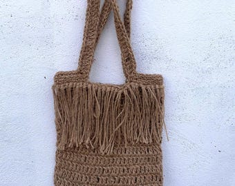 May include: A brown jute tote bag with fringe detail. The bag has two woven handles and a rectangular body. The fringe is at the top of the bag, adding a decorative element. The bag is hanging against a white wall.