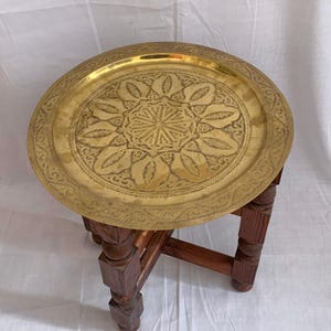 Pode incluir: Uma bandeja redonda, cor de latão, com intrincados padrões florais gravados, repousa sobre uma pequena mesa de madeira escura. O design da bandeja apresenta uma estrela central e formas semelhantes a folhas. A mesa tem pernas quadradas.